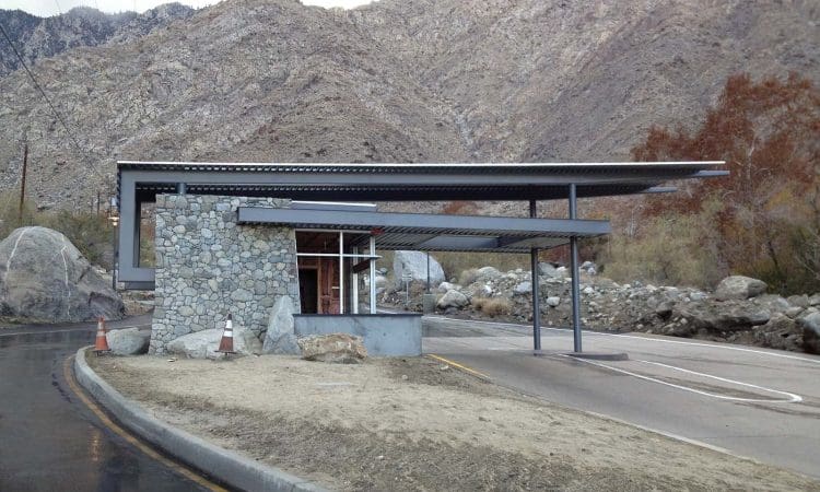 A modern stone and metal toll booth stands beside a curved road, with mountains and rocky terrain in the background.