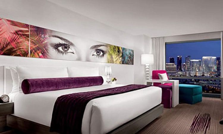 Modern hotel room with a large bed, purple accents, wall art of eyes above the headboard, seating area, and a window view of a city skyline at night.