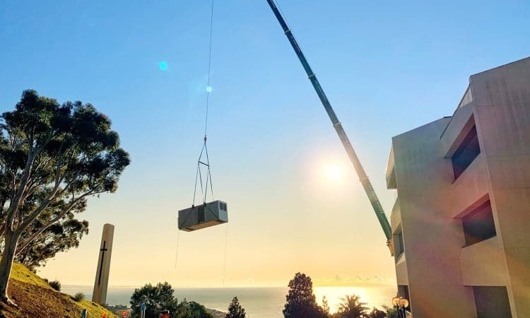 A large crane lifts an industrial HVAC unit near a multi-story building at sunset, with trees and the ocean visible in the background.