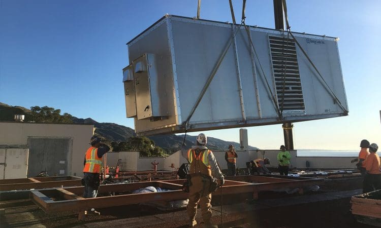 Construction workers guide a large HVAC unit being lifted by a crane onto a building rooftop under clear sky.