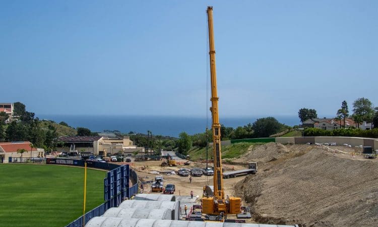 A large yellow crane operates at a construction site near a grassy field, with the ocean and several buildings visible in the background under a clear sky.