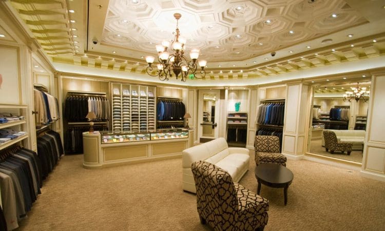 Luxury men's clothing store interior with suits, shirts, and ties displayed along the walls, a sofa and chairs in the center, and ornate ceiling lighting above.