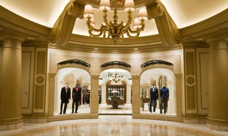 A chandelier-lit hallway leads to a formal menswear store with mannequins in suits and shirts displayed in the windows.