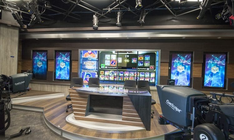 A modern TV studio set with a news desk, cameras, overhead lights, and large video screens displaying gaming graphics in the background.