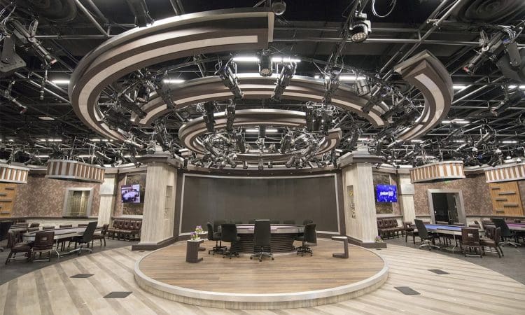 A modern television studio with a central round stage, multiple chairs, overhead lighting rigs, and surrounding tables; monitors and equipment are visible throughout the space.