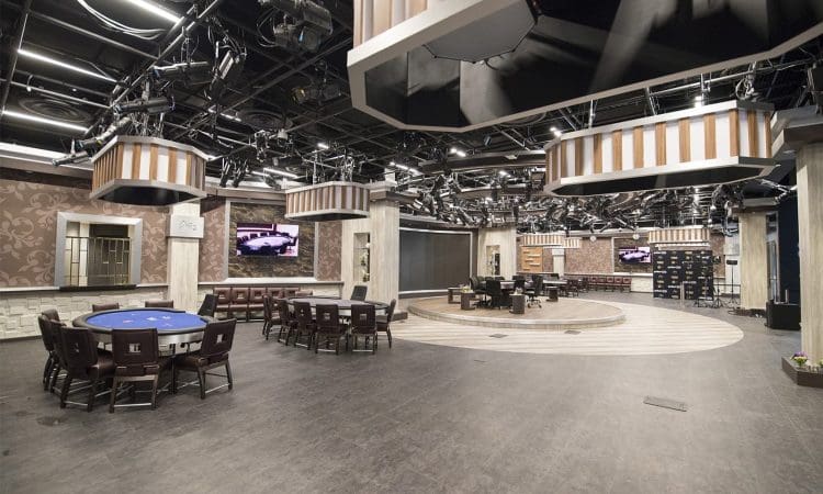 Spacious, well-lit poker room with multiple poker tables, cushioned chairs, large TV screens, and decorative wall panels under a complex ceiling with stage lighting.