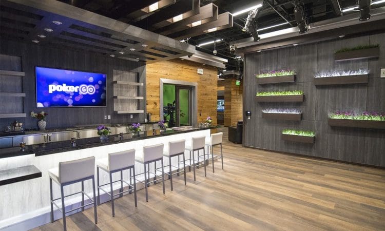 Modern lounge with a bar, five white bar stools, a wall-mounted TV displaying "pokerGO," decorative plants, and wood floors under studio lighting.