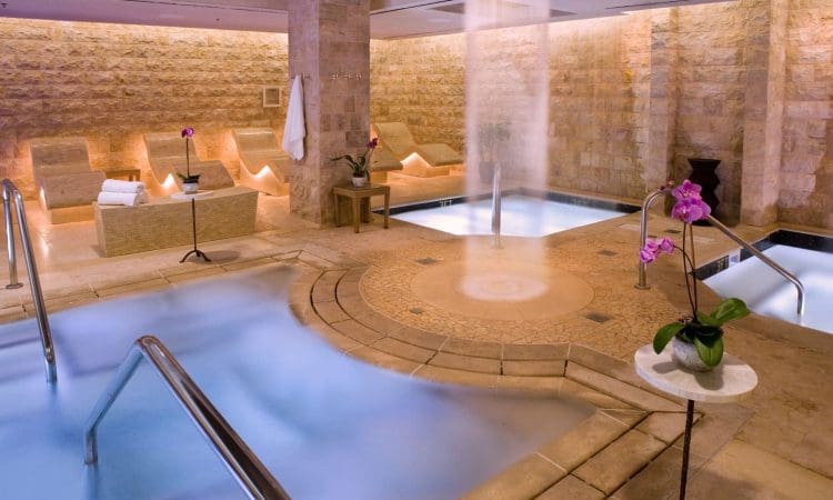 Indoor spa area with stone walls, mist rising from hot tubs, lounge chairs with towels, potted orchids, and soft ambient lighting.