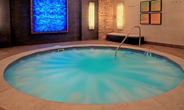 Round indoor hot tub with bubbling water, tiled edge, and metal handrail in a modern spa room with stone and art decor on the walls.