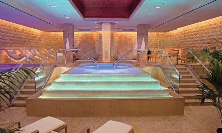 Indoor spa area with illuminated central pool, stairs with glass railings, stone walls, and ambient colored lighting; lounge chairs and plants in the foreground.