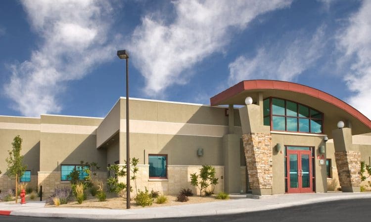 Single-story, modern building with stone accents, large windows, red double doors, and a curved roofline, set against a blue sky with scattered clouds.