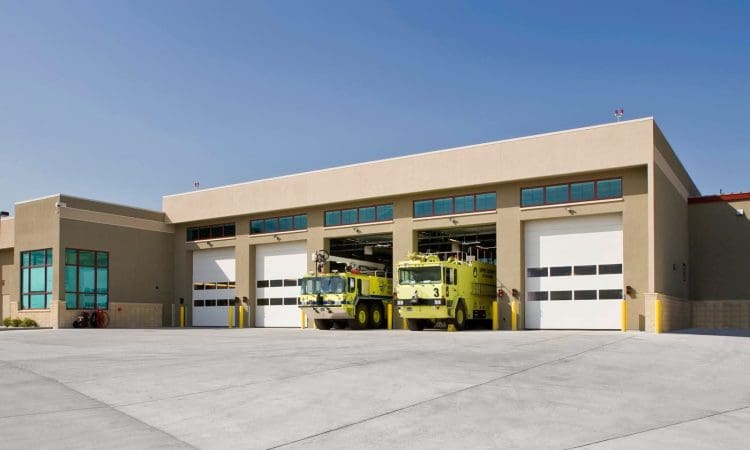 A modern fire station building with five garage bays, two of which contain yellow fire trucks parked inside.