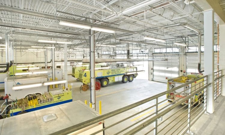 Interior view of a bright, spacious fire station bay with multiple large yellow fire trucks parked inside, seen from an upper walkway.