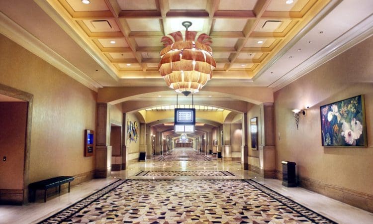 Wide hotel hallway with patterned floor, ornate ceiling light fixture, wall art, benches, and a digital sign suspended from the ceiling at the center.