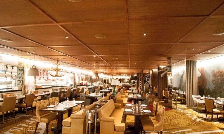Spacious, well-lit restaurant interior with tan upholstered chairs, set tables, patterned carpet, and an open kitchen area in the background.