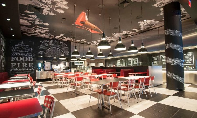 Modern restaurant interior with red and white chairs, black tables, menu boards on the wall, and decorative ceiling art. The space appears clean and well-lit, with no people present.