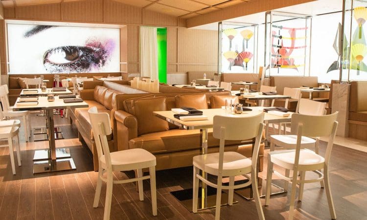 Modern restaurant interior with tan leather couches, white chairs, set tables, wood flooring, and large contemporary artwork on the walls.
