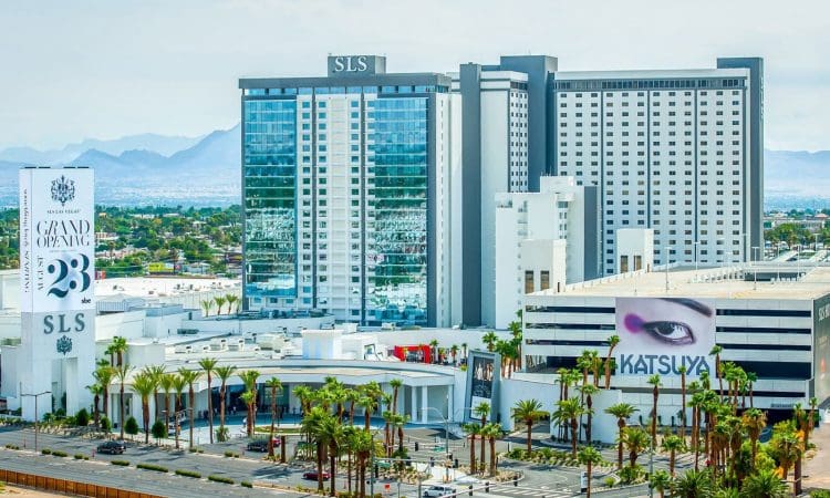 A view of the SLS hotel and casino complex in Las Vegas, featuring modern high-rise buildings, palm trees, and billboards, with mountains visible in the background.