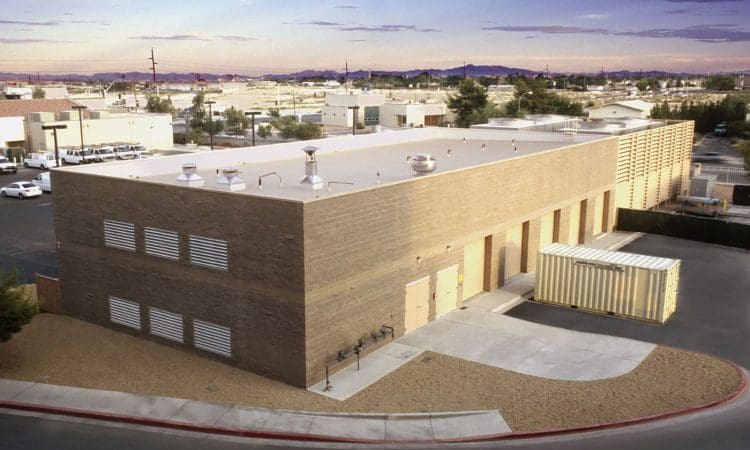 A large, rectangular industrial building with a flat roof and ventilation systems is shown in a suburban area, with a shipping container placed nearby and desert landscaping surrounding the property.