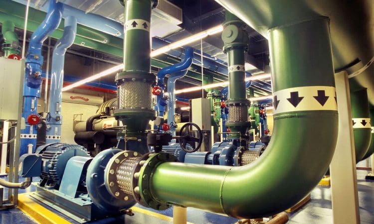 Large industrial green and blue pipes with directional arrow labels, mechanical equipment, and valves inside a brightly lit facility.