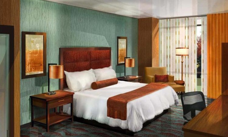 Modern hotel room with a king-sized bed, two bedside tables with lamps, an armchair, a desk with a chair, and large windows with sheer curtains.