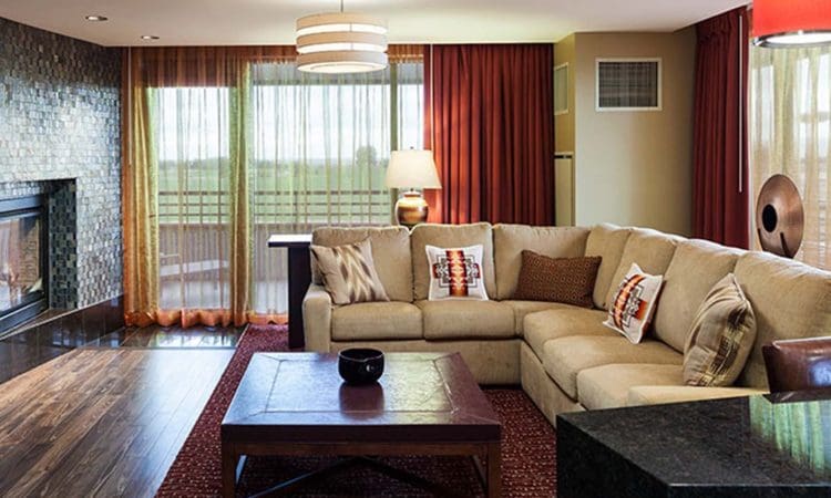 Modern living room with a beige sectional sofa, accent pillows, wooden coffee table, fireplace, floor lamp, and large windows with sheer and red curtains.