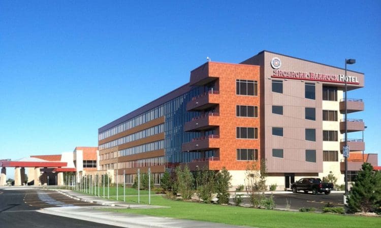 A modern, multi-story hotel building with red and beige exterior, large windows, and a parking lot, under a clear blue sky.