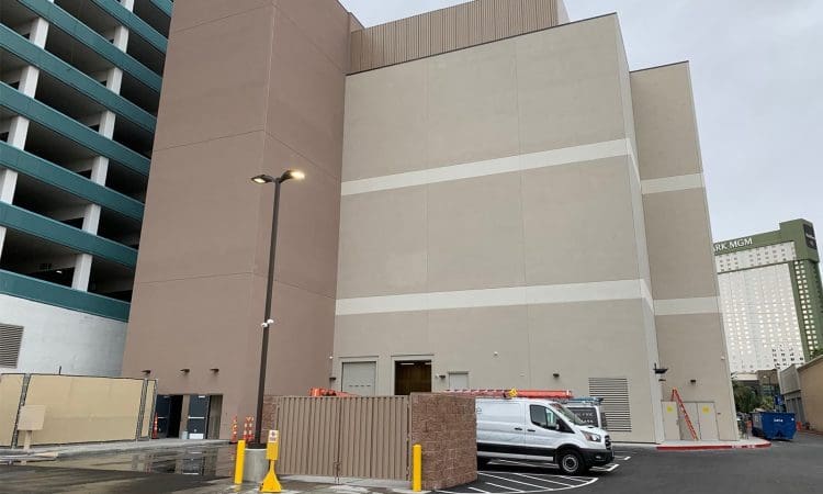 A large beige utility building with a white van parked outside, adjacent to a multi-level parking structure, under an overcast sky.