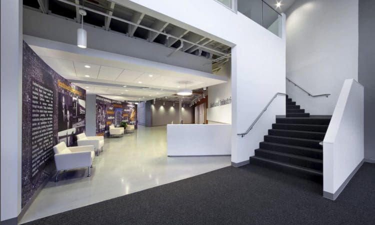 Modern office lobby with white chairs, wall murals, a black carpeted staircase, and overhead lighting. The space appears open and well-lit with clean architectural lines.