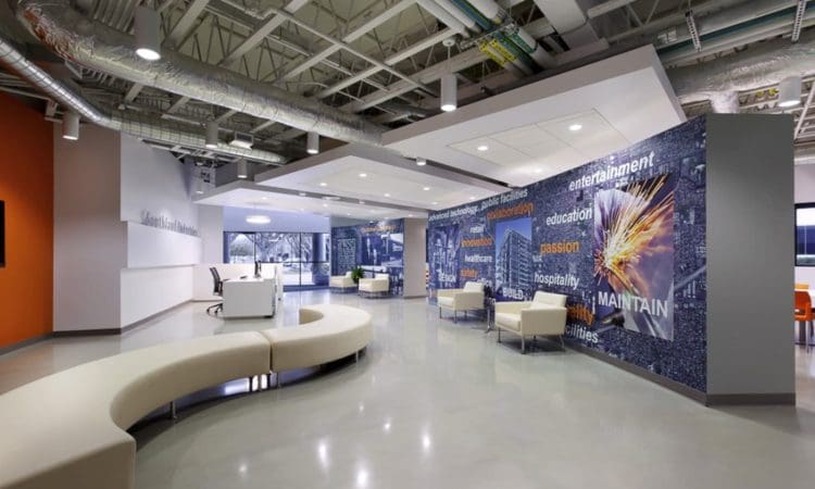 Modern office lobby with curved seating, white chairs, and a mural wall featuring industry words and abstract graphics. Exposed ceiling pipes and neutral tones dominate the space.