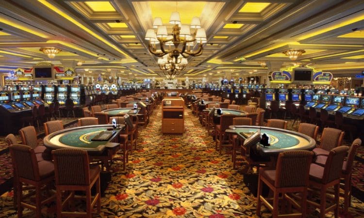Large, brightly lit casino floor with rows of slot machines, card tables, and patterned carpet under an ornate ceiling with chandeliers.