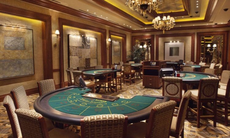 Casino gaming room with several empty card tables, upholstered chairs, decorative walls, and chandeliers under warm lighting.