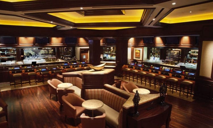 A spacious, upscale bar with wooden floors, plush seating, a long counter lined with barstools, and warm, ambient lighting.