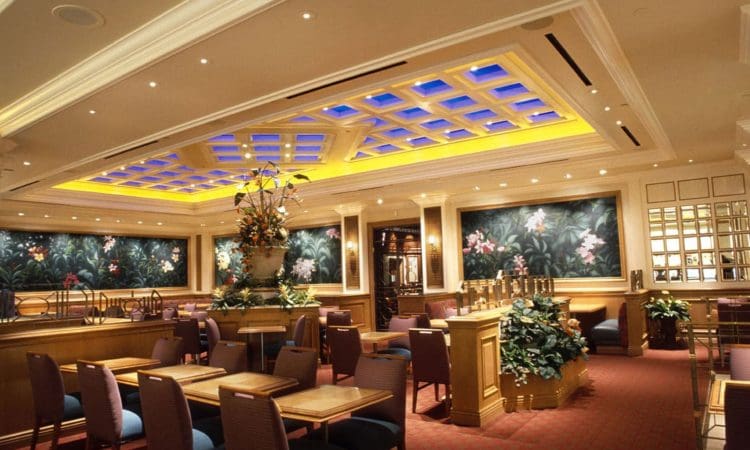 Upscale restaurant interior with red carpet, wooden tables and chairs, floral wall art, decorative plants, and a ceiling with blue and yellow lighting accents.