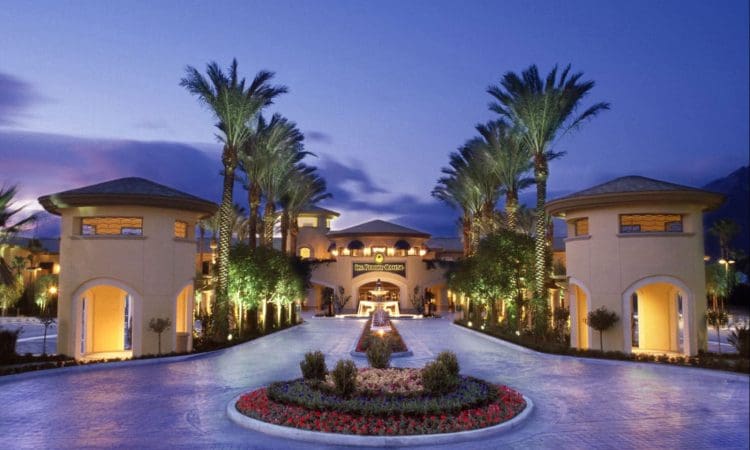A luxury resort entrance with palm trees, landscaped gardens, round pavilions, and illuminated pathways at dusk.