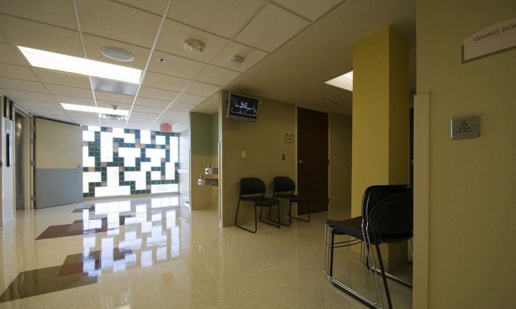 Empty hallway in a medical facility with chairs along the wall, a television mounted near the ceiling, and patterned glass windows at the end of the corridor.
