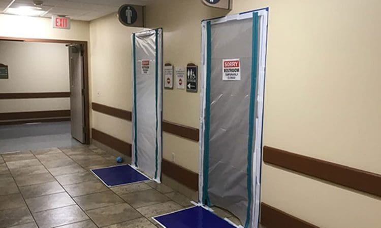 Two restroom doors in a hallway are covered with plastic sheets and “Sorry! Restroom temporarily out of service” signs. Blue mats are placed on the floor in front of each door.
