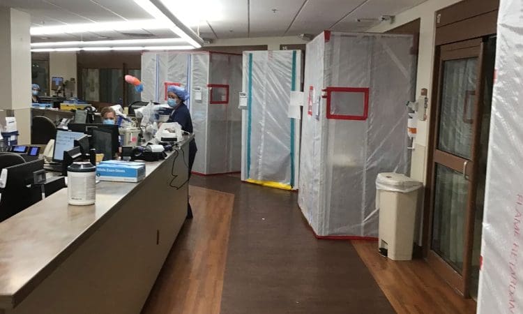 Hospital hallway with plastic containment barriers and healthcare workers at desks, likely indicating infection control or construction in progress.