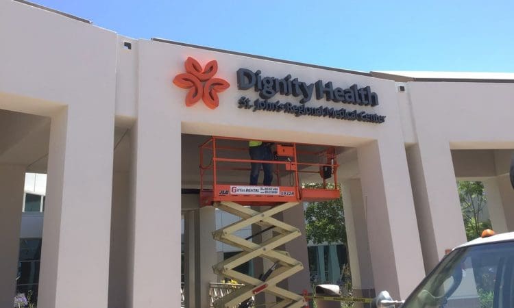 A worker on a scissor lift installs or repairs the Dignity Health St. Louise Regional Medical Center sign on a building exterior.