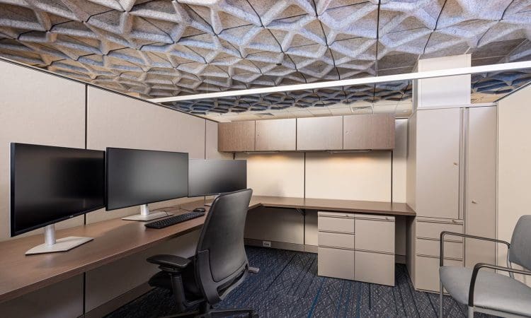 Modern office cubicle with four large computer monitors, a keyboard, gray office chairs, overhead cabinets, and geometric patterned ceiling tiles.