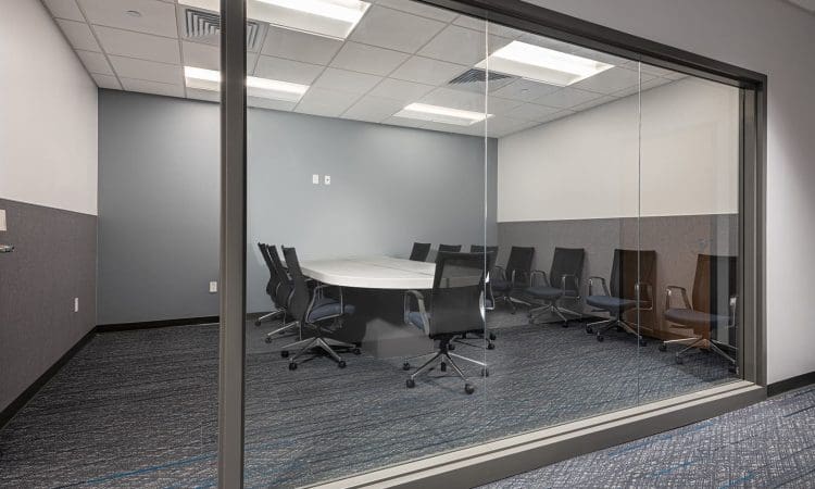 Glass-walled conference room with a round table and ten black office chairs arranged around it, viewed from outside the room.