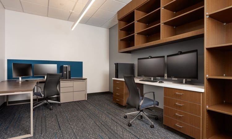 Modern office with two desks, each equipped with computer monitors and office chairs, wood shelving units, and a carpeted floor.