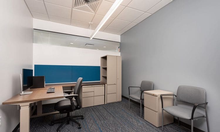 A modern office with a desk, computer, office chair, storage cabinets, and two guest chairs against a blue accent wall.