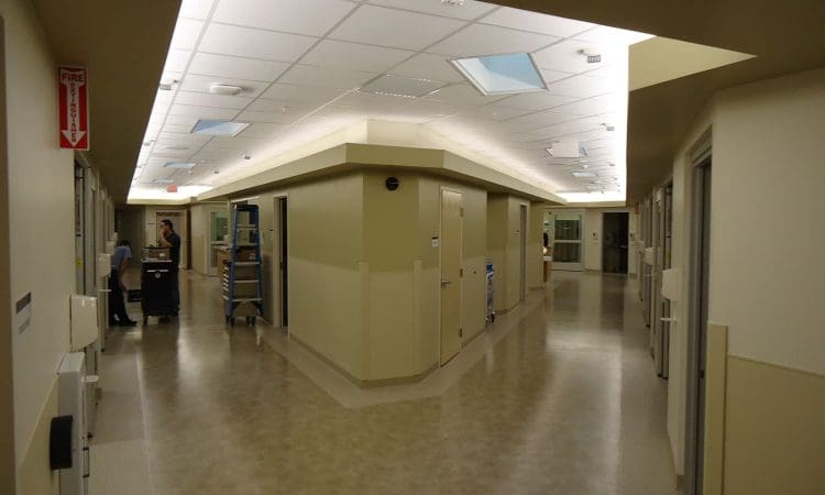 Hospital hallway with beige floors and walls, overhead fluorescent lighting, and several doors leading to patient rooms; two people and equipment carts are visible in the background.