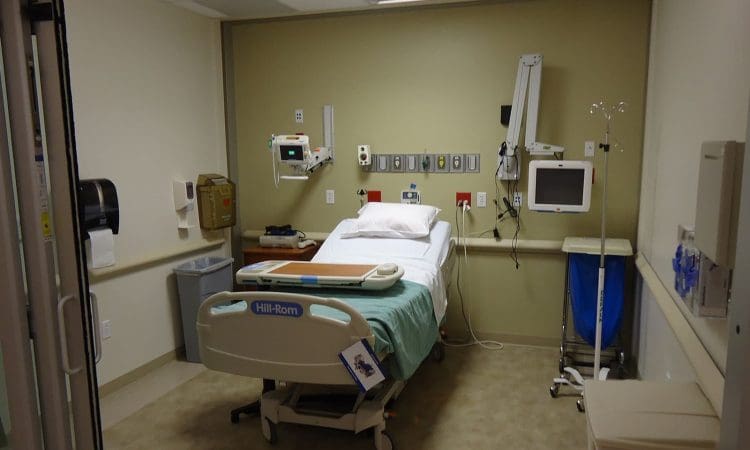 A hospital room with a bed, medical equipment, monitor, and supplies against a beige wall, viewed from the entrance.