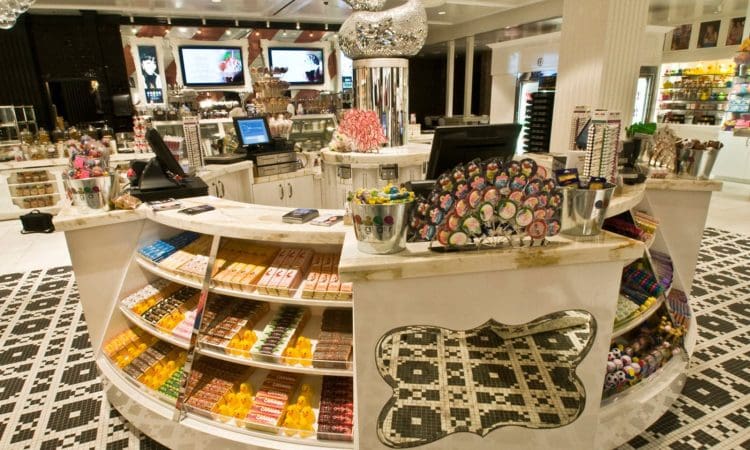 A brightly lit candy store interior features curved display shelves filled with assorted colorful candies and lollipops, with two registers on the counter.