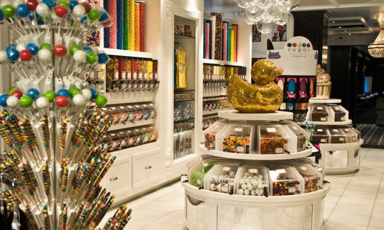 A brightly lit candy store features colorful lollipops, candy dispensers, and a large golden duck centerpiece on a round display.