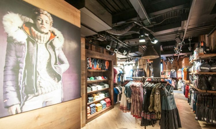 Clothing store interior with racks of clothes, shelves of folded items, hats, and a large poster of a person wearing a winter jacket on the left wall.
