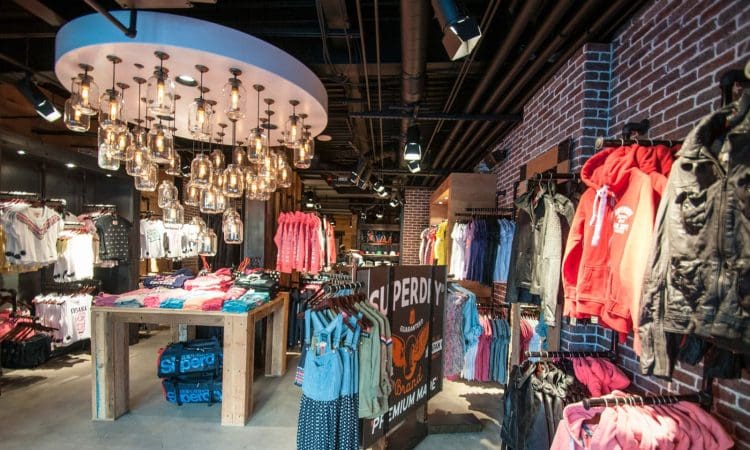 Clothing store interior with racks of assorted clothes, folded shirts on tables, exposed brick walls, and hanging glass light fixtures.