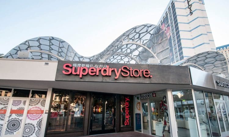 Exterior of a Superdry Store with a modern, wavy glass canopy and neighboring shops, located in an urban area with a tall building in the background.